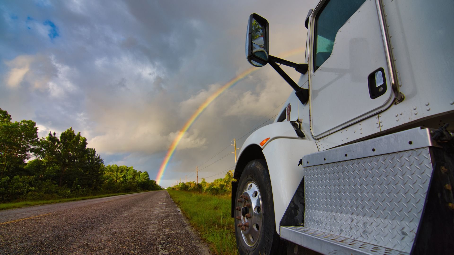 White truck parked on rural road with vibrant rainbow in cloudy sky